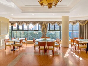 A spacious dining area with round tables and chairs, large windows with curtains, and a chandelier.