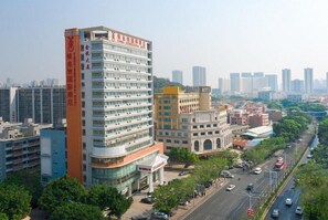 A multi-story hotel with a prominent orange vertical sign, a modern building with a glass facade, and a street view with cars and trees.