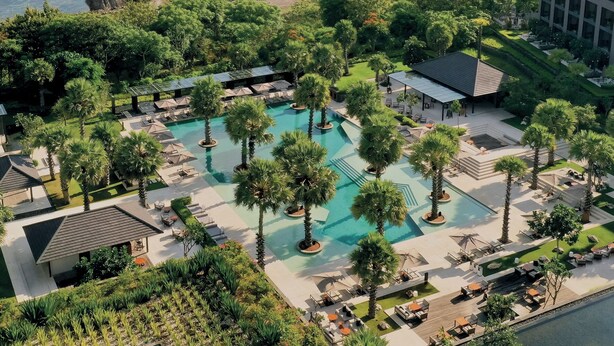 Vue aérienne d’un complexe hôtelier avec une piscine centrale entourée de palmiers et d’espaces de détente.