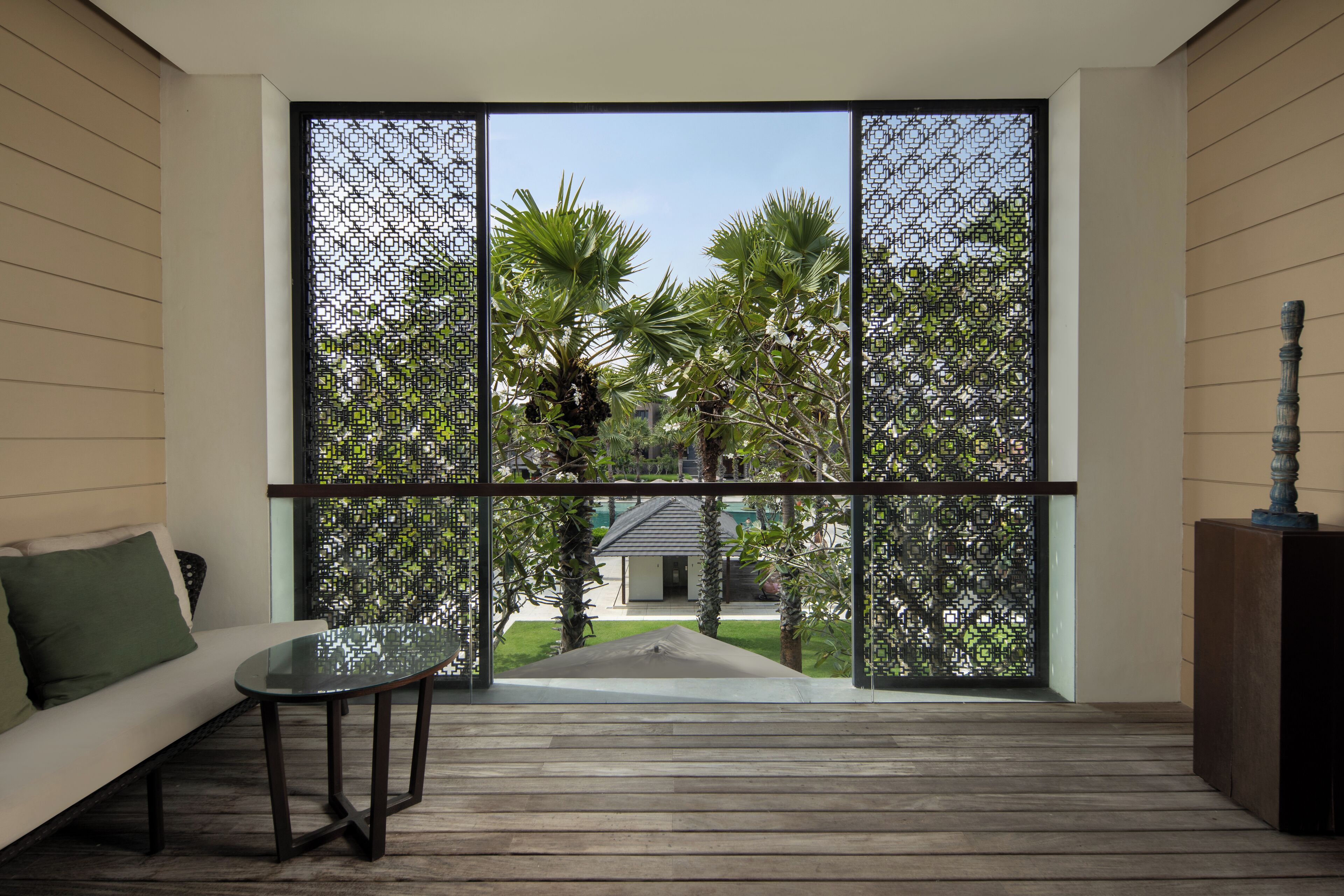 Una habitación con suelo de madera, una mesa de vidrio, un sofá blanco con un cojín verde, y vistas a través de una ventana con marco negro y un diseño geométrico.