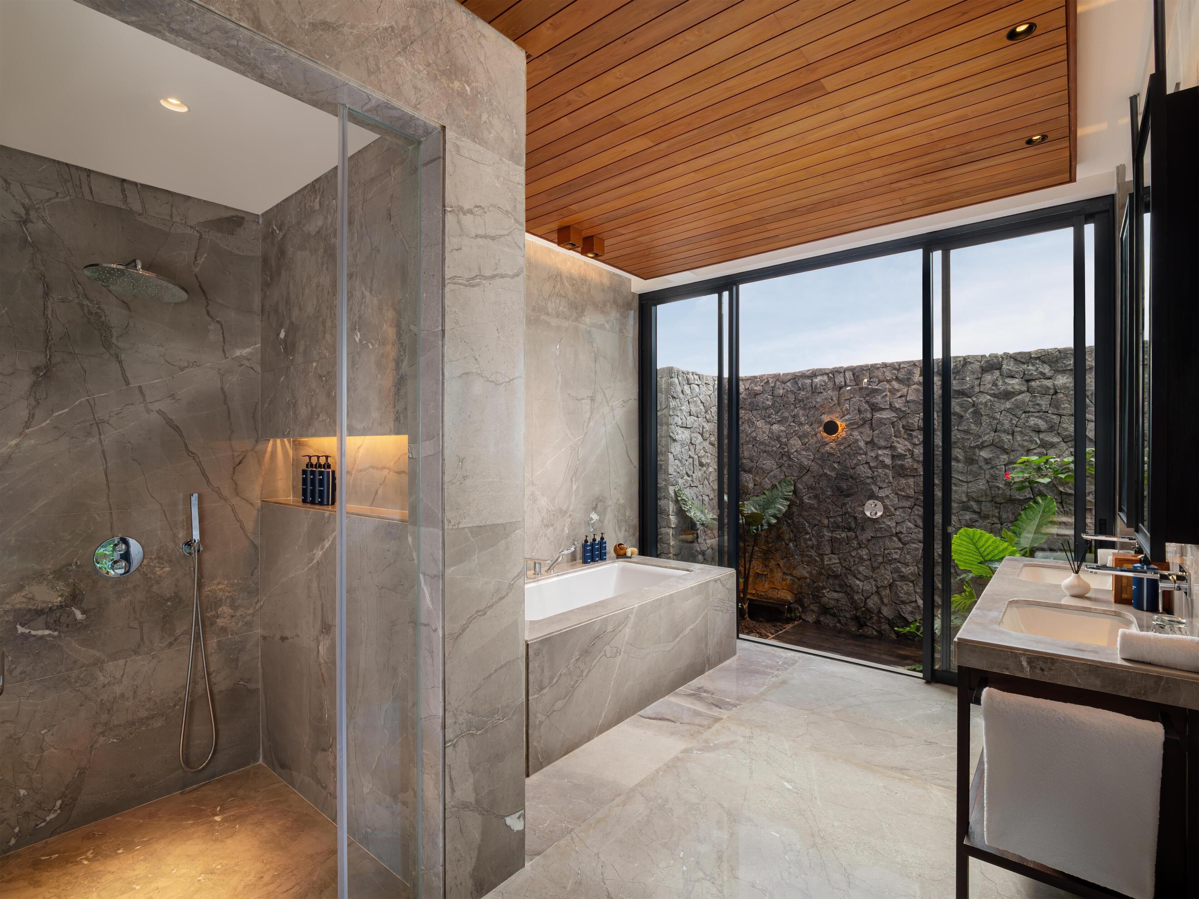 A modern bathroom with a glass-enclosed shower, a bathtub, and a large window offering a view of a stone wall and greenery.