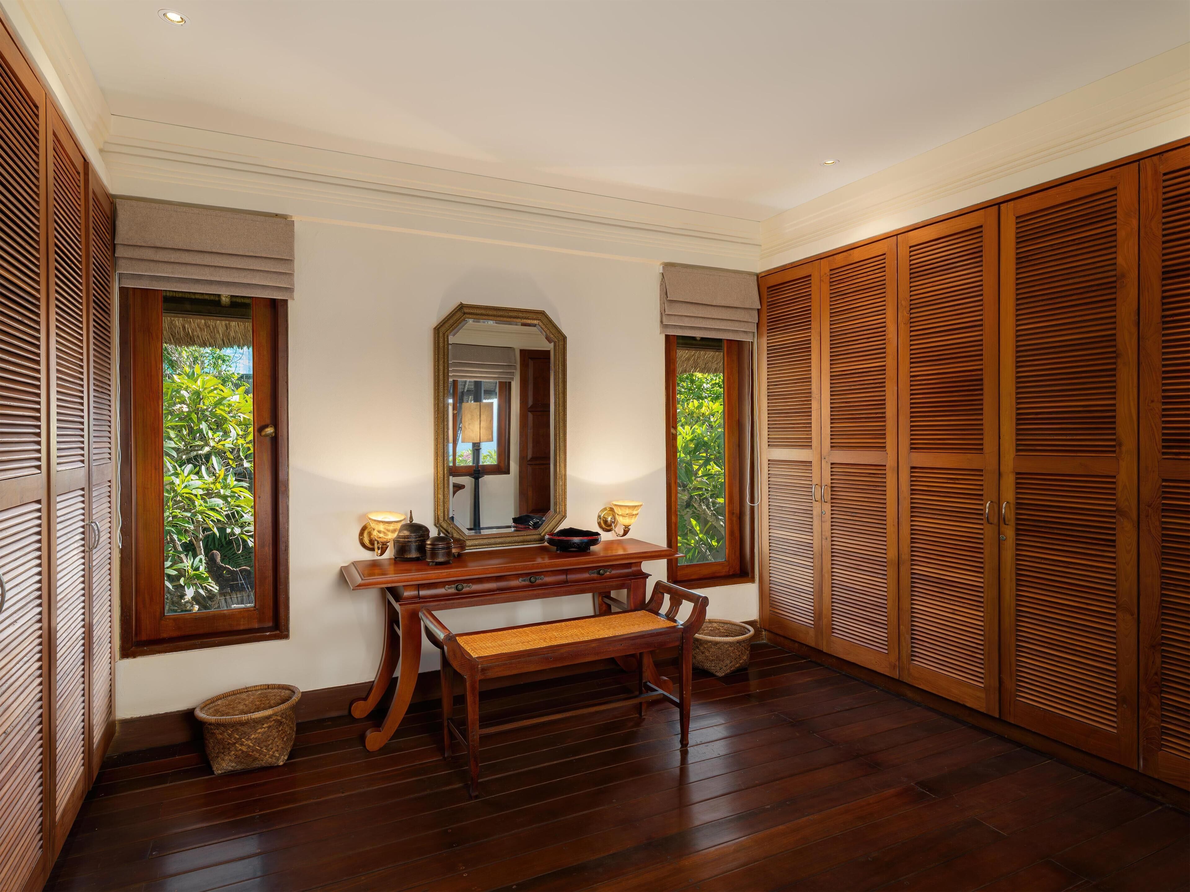 A room with a wooden bench, a table, a mirror, and wooden louvered closets.