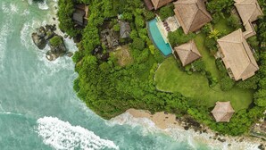 Vue aérienne d’une propriété située sur la côte, avec une piscine, des constructions à toits de chaume et une végétation luxuriante.