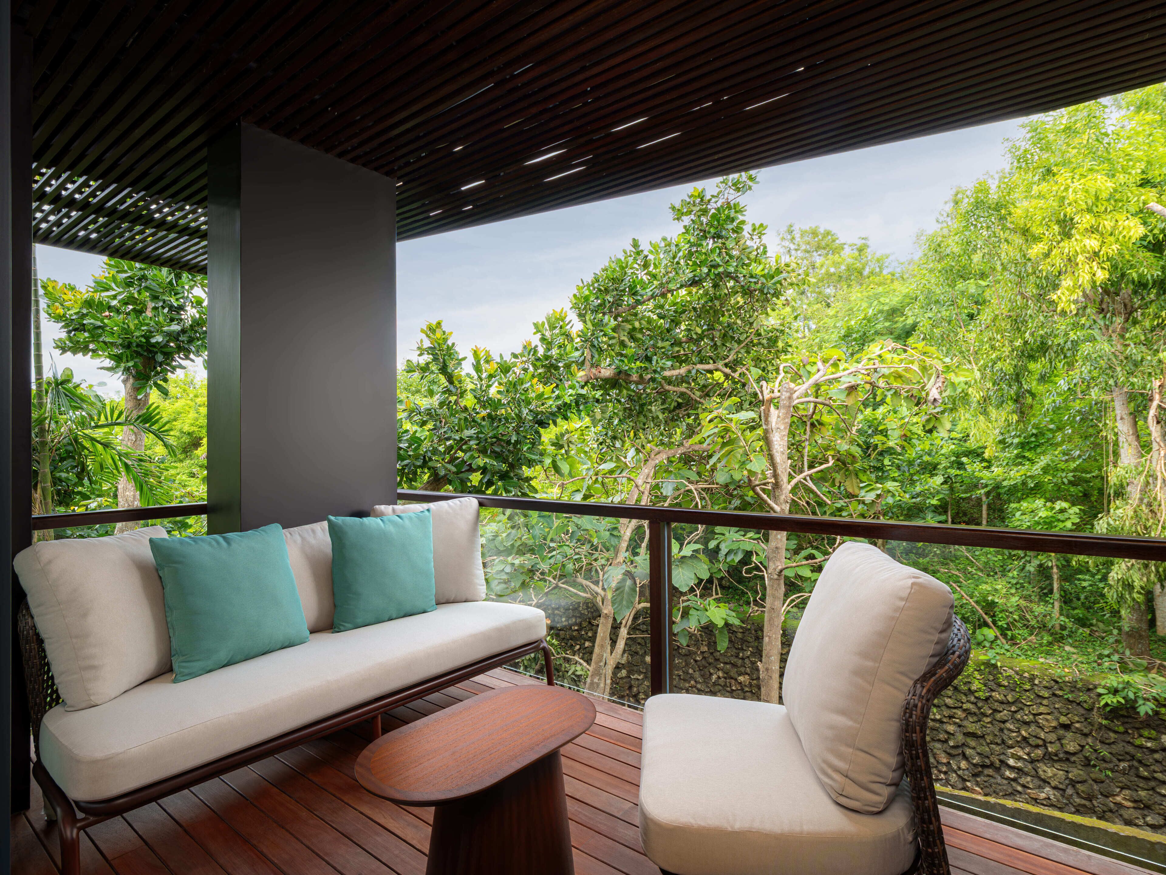 Un balcón con sofá y sillón, una mesa de madera y vistas a un follaje exuberante.