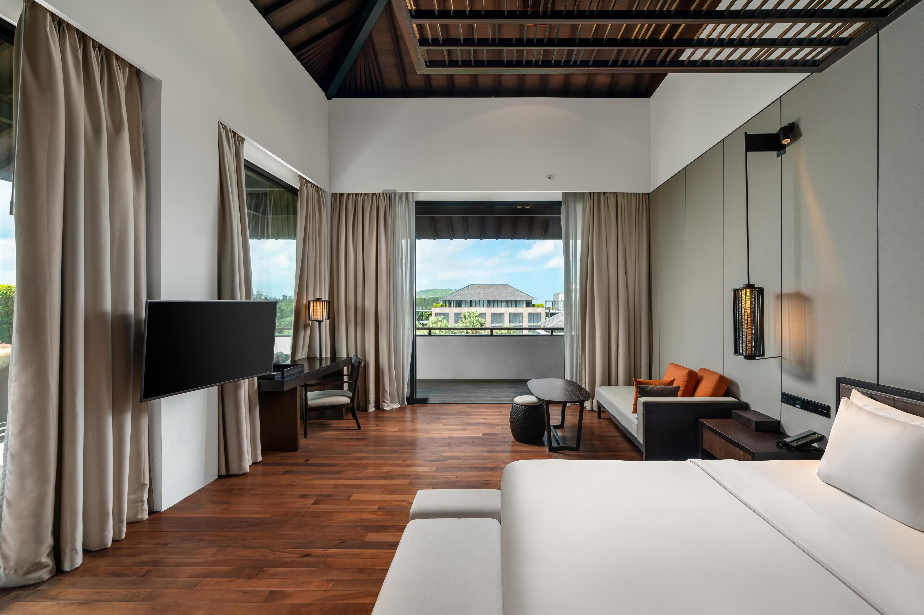 A modern hotel room with a large bed, a desk, a sofa, and a TV. The room has wooden flooring and large windows with curtains.
