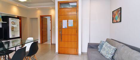 Una sala moderna con un sofá gris, una mesa de comedor de cristal con sillas, una puerta de madera con un aviso y una alfombra geométrica.