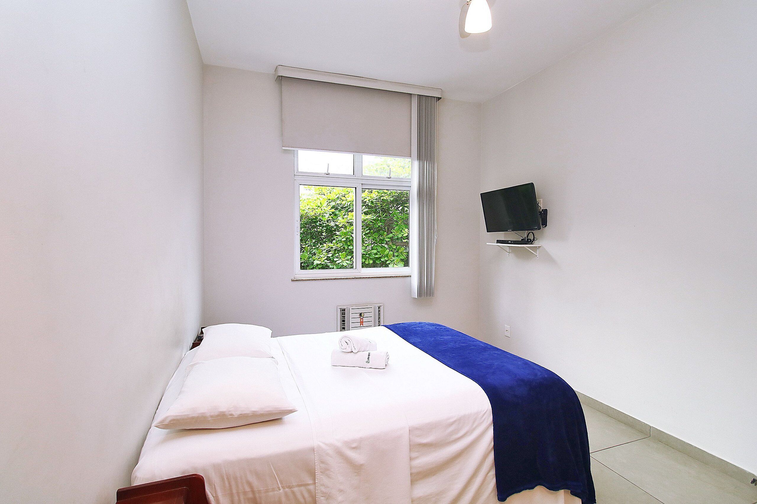 Un dormitorio con cama, televisor montado en la pared y ventana con vistas a la vegetación.