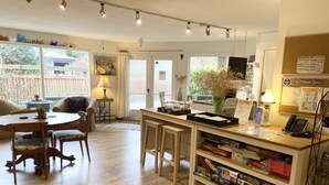 A cozy living area with a wooden dining table, comfortable seating, a small kitchen area with a coffee machine, and a bookshelf filled with board games. - Wisteria Guest House (Salt Spring Island)