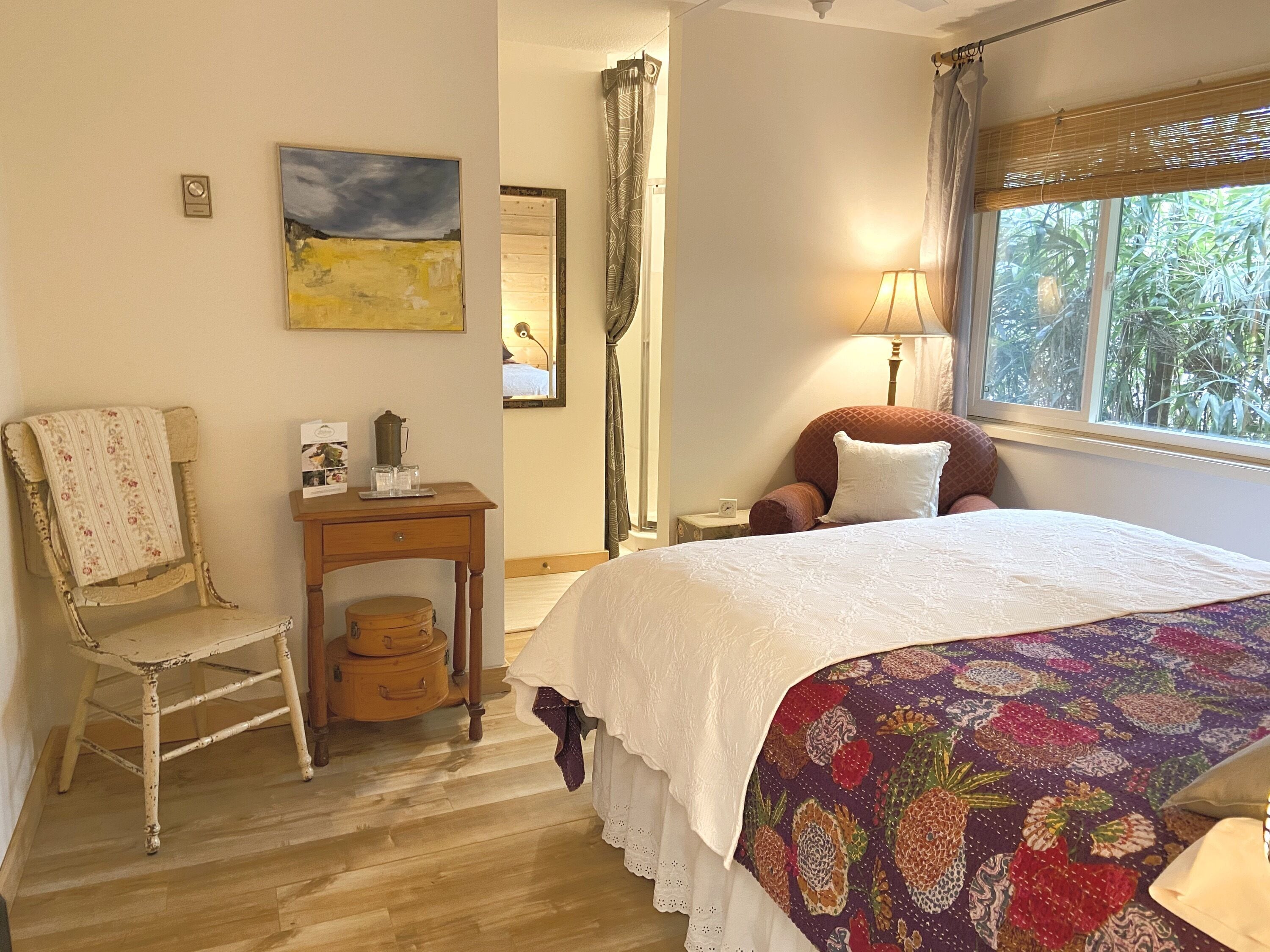 A bedroom with a bed, a chair, a small table with a lamp, a window with a view of greenery, and artwork on the walls.