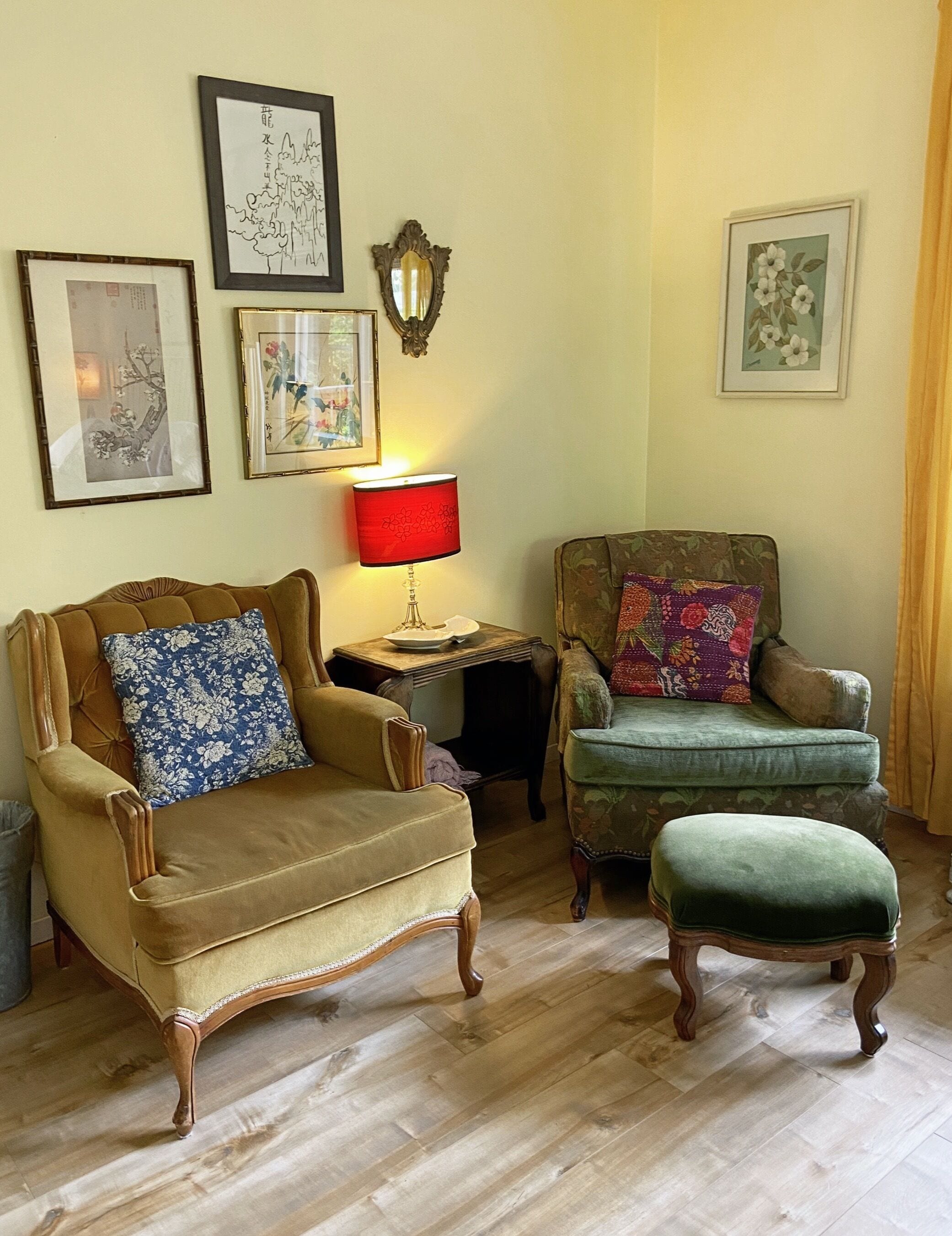 Un salon traditionnel avec un fauteuil marron, un fauteuil vert et un pouf assorti. On y trouve une table avec une lampe rouge, ainsi que plusieurs œuvres d’art encadrées accrochées aux murs.