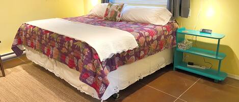 A bed with a colorful quilt, a bedside table with a phone, and a window with curtains.