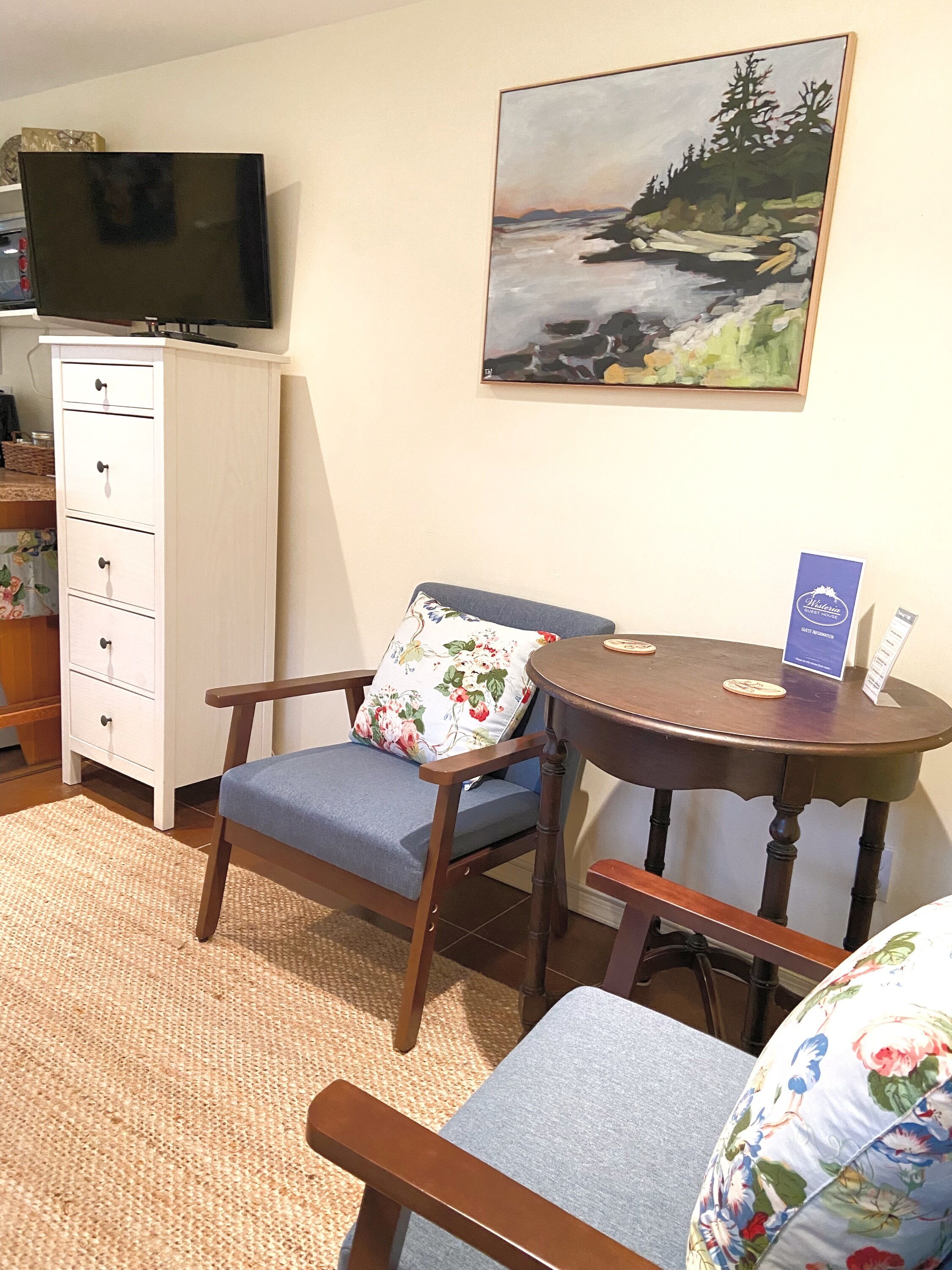 Ein Zimmer mit Fernseher auf einer Kommode, einem Bild an der Wand, einem Tisch mit Untersetzern und einem Sessel mit einem blumigen Kissen.