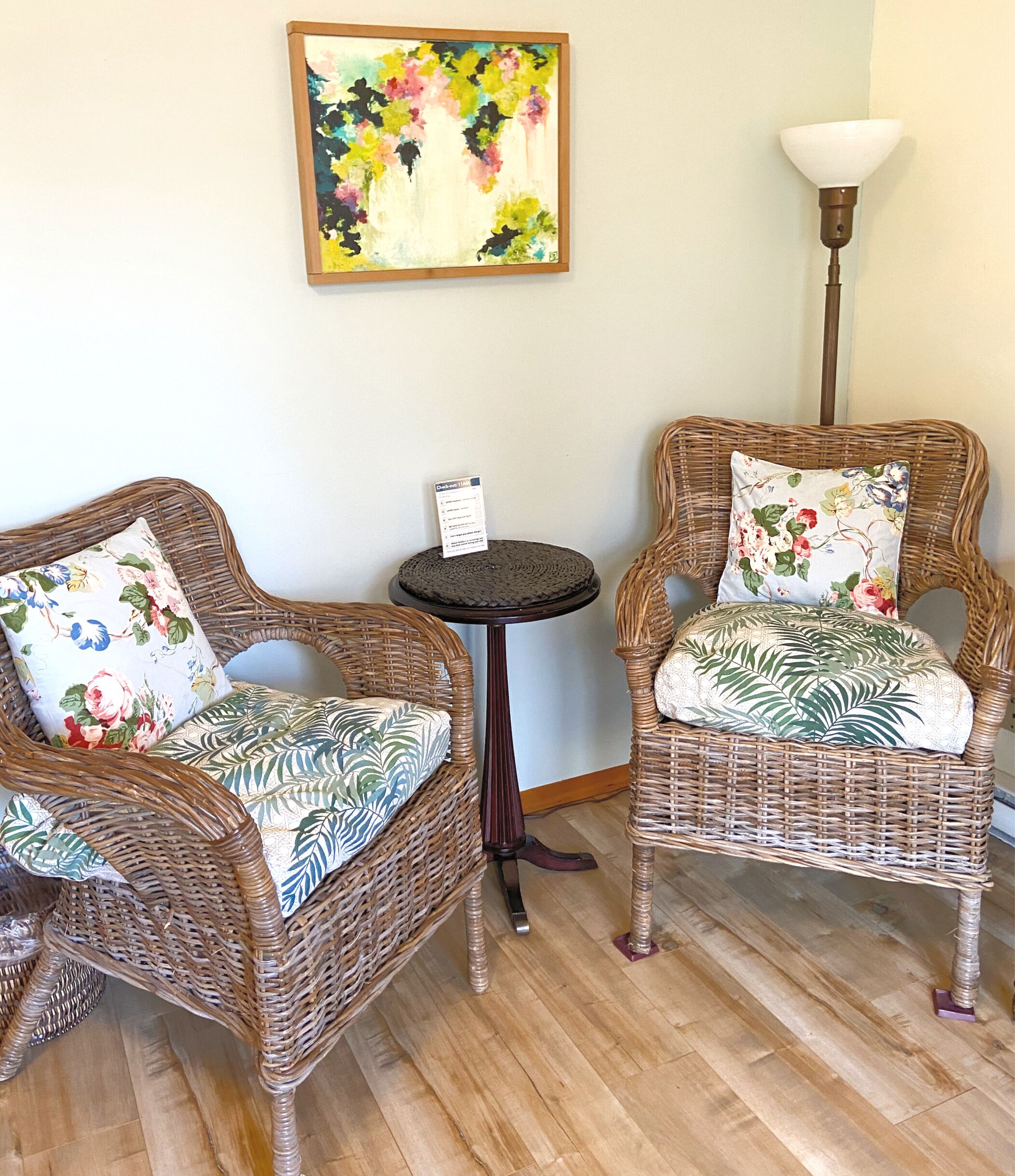 Two wicker chairs with floral cushions, a small round table with a sign, and a floor lamp.