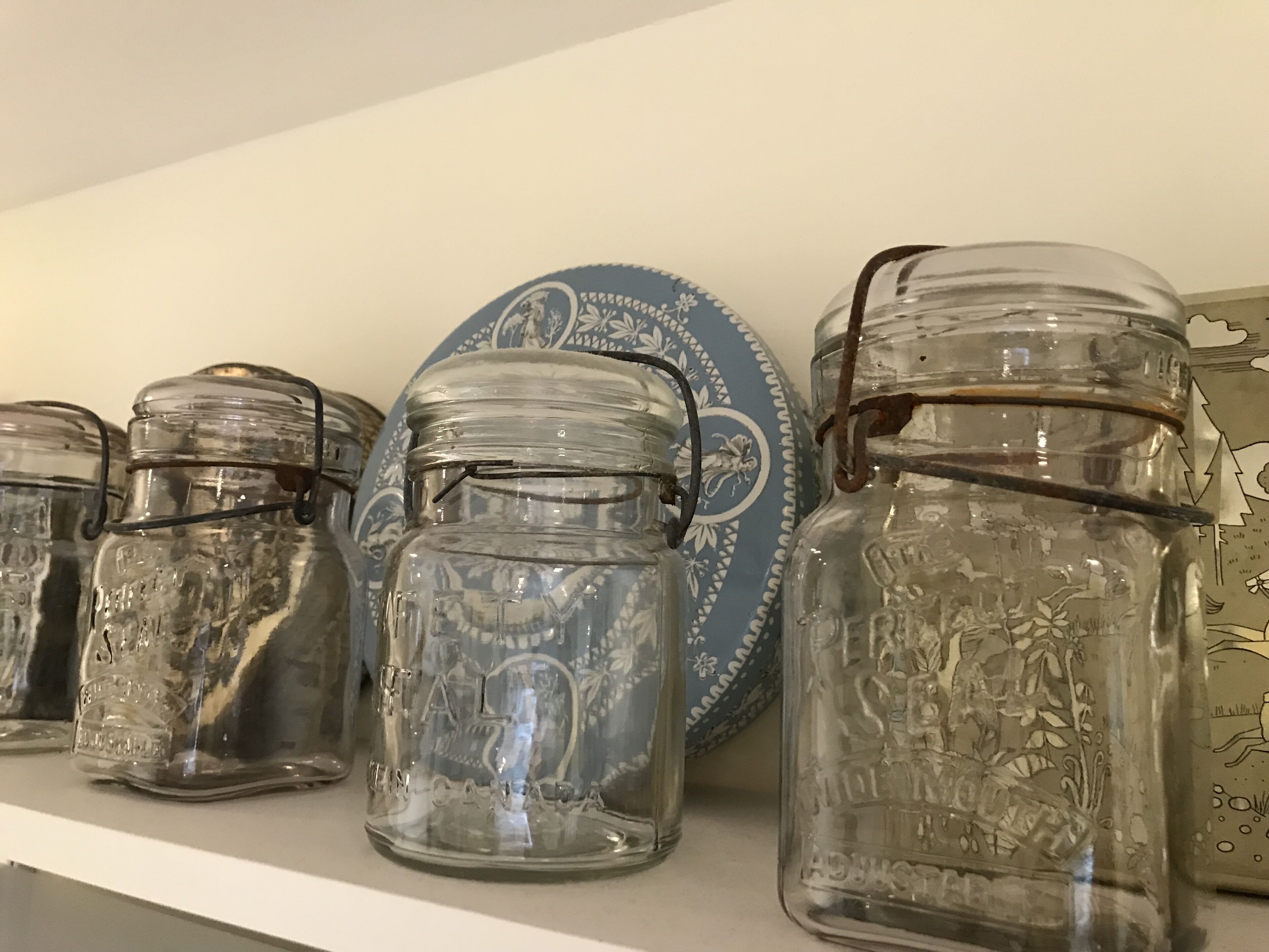 A shelf with four glass jars, each with metal clasps, and a decorative plate with a floral pattern.