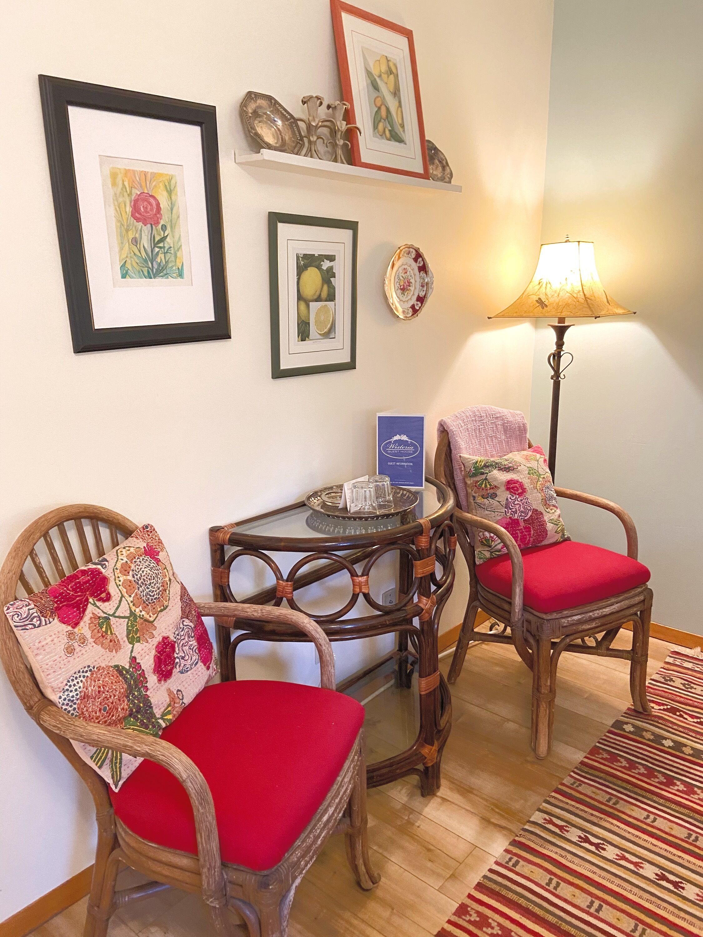 A room with two wooden chairs with red cushions, a small table with a tray, and a lamp. There are framed pictures on the wall and a patterned rug on the floor.