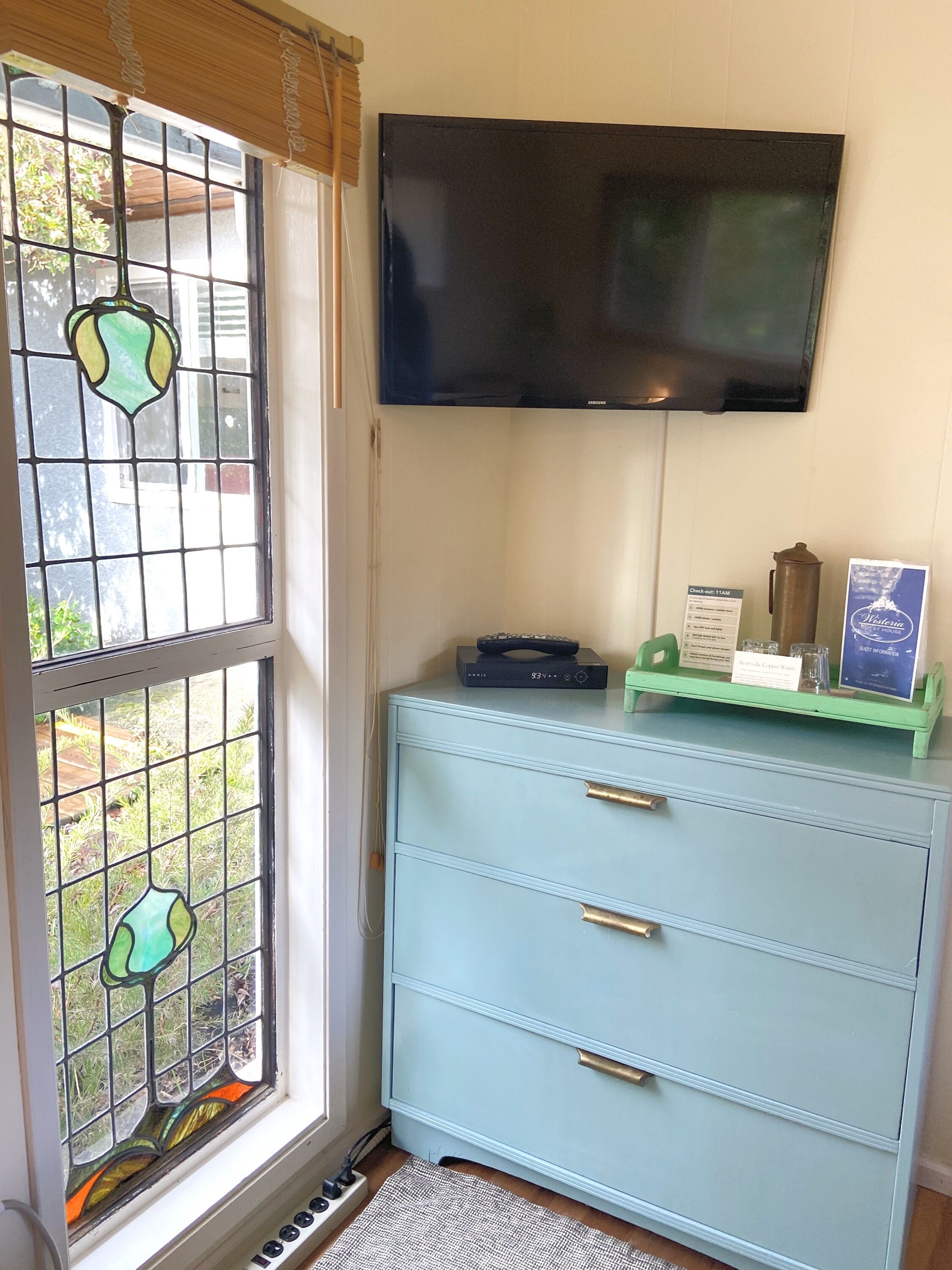Una habitación con un armario azul claro, un televisor de pantalla plana montado en la pared y un vitral con diseño verde y amarillo.