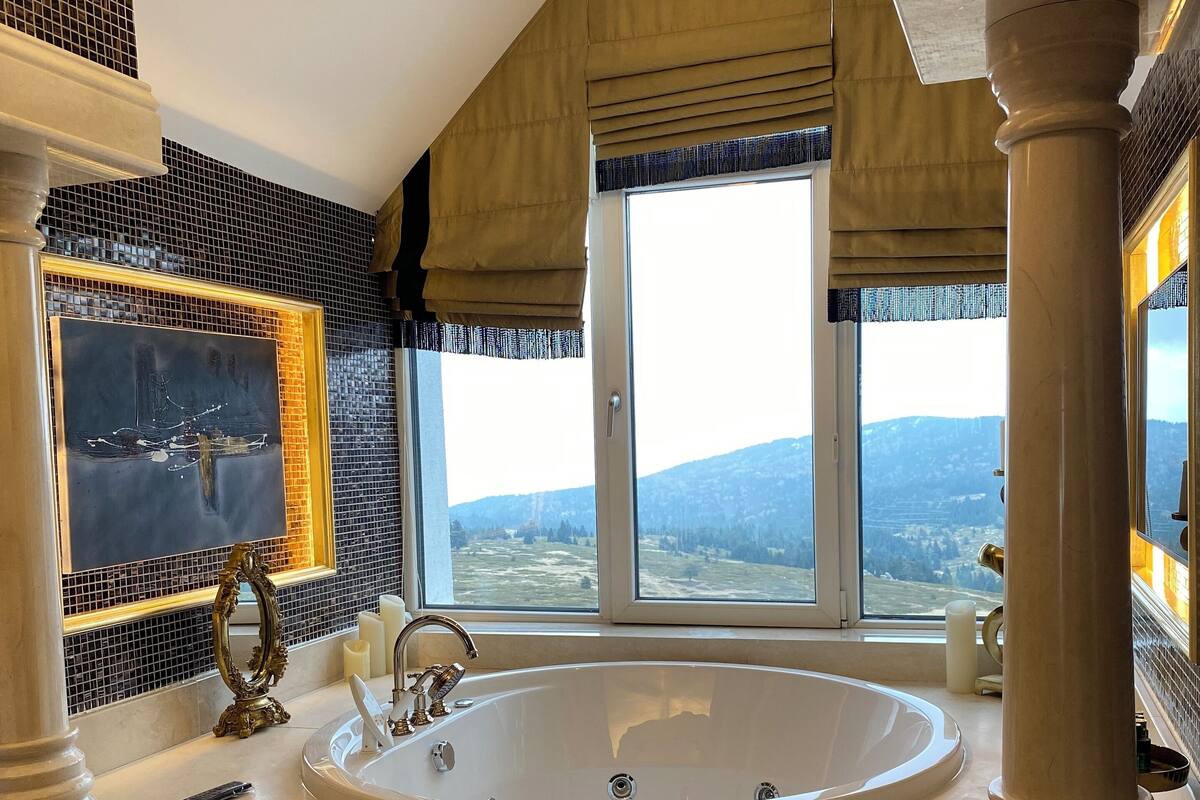 A spacious bathroom with a large bathtub, a mirror, and a view of the mountains through large windows.