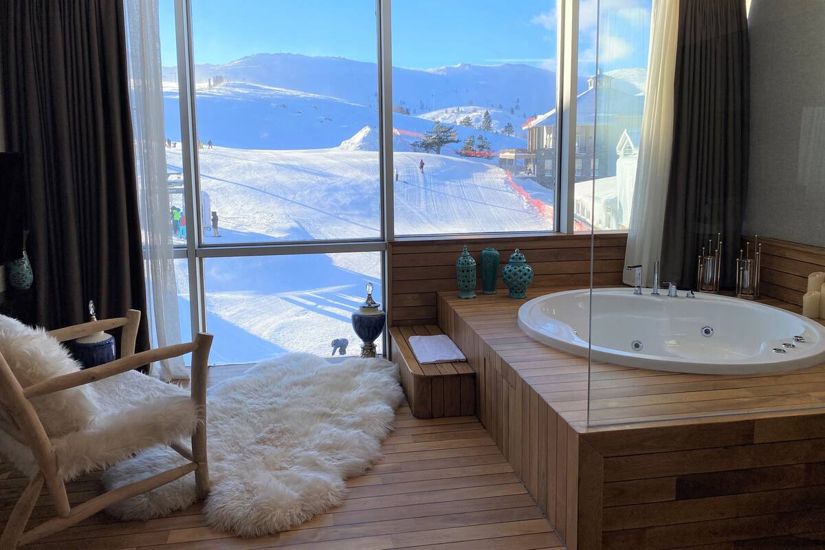 A modern bathroom with a large window, wooden flooring, a white fur rug, a wooden chair, a bathtub, and decorative vases.