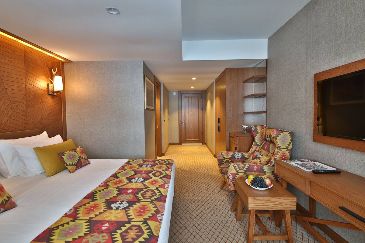 A hotel room with a bed, a desk, a chair, a TV, and a small table with fruit.
