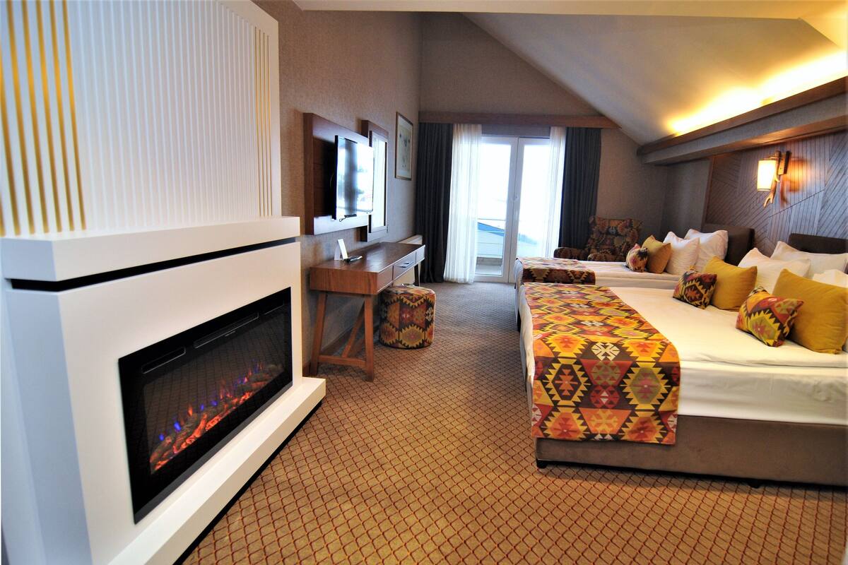 A hotel room with a fireplace, a desk, two beds with patterned bedding, and a view of the outdoors.