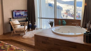 Une salle de bain moderne avec une grande baignoire, un plancher en bois, un fauteuil moelleux et un téléviseur affichant l’écran d’accueil de l’hôtel.