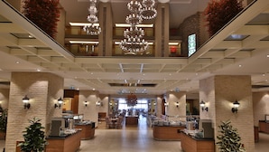 Un hall d’hôtel spacieux, doté de plusieurs espaces salon, d’un escalier central et d’un lustre moderne.