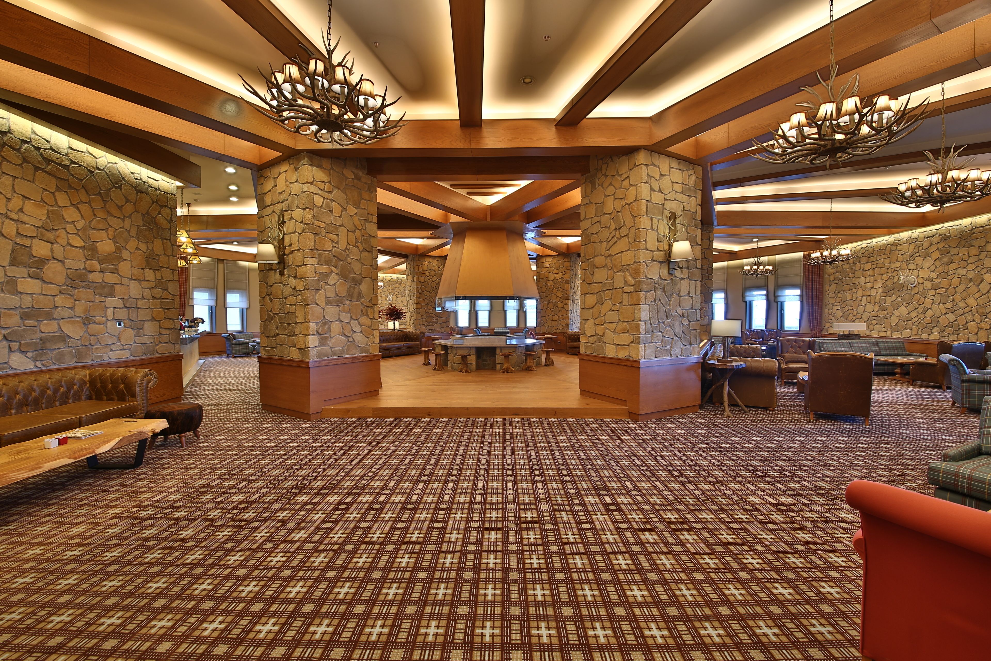 Un hall espacioso con alfombra estampada, paredes de piedra, vigas de madera y varias zonas de estar, incluyendo una mesa larga y sofás.