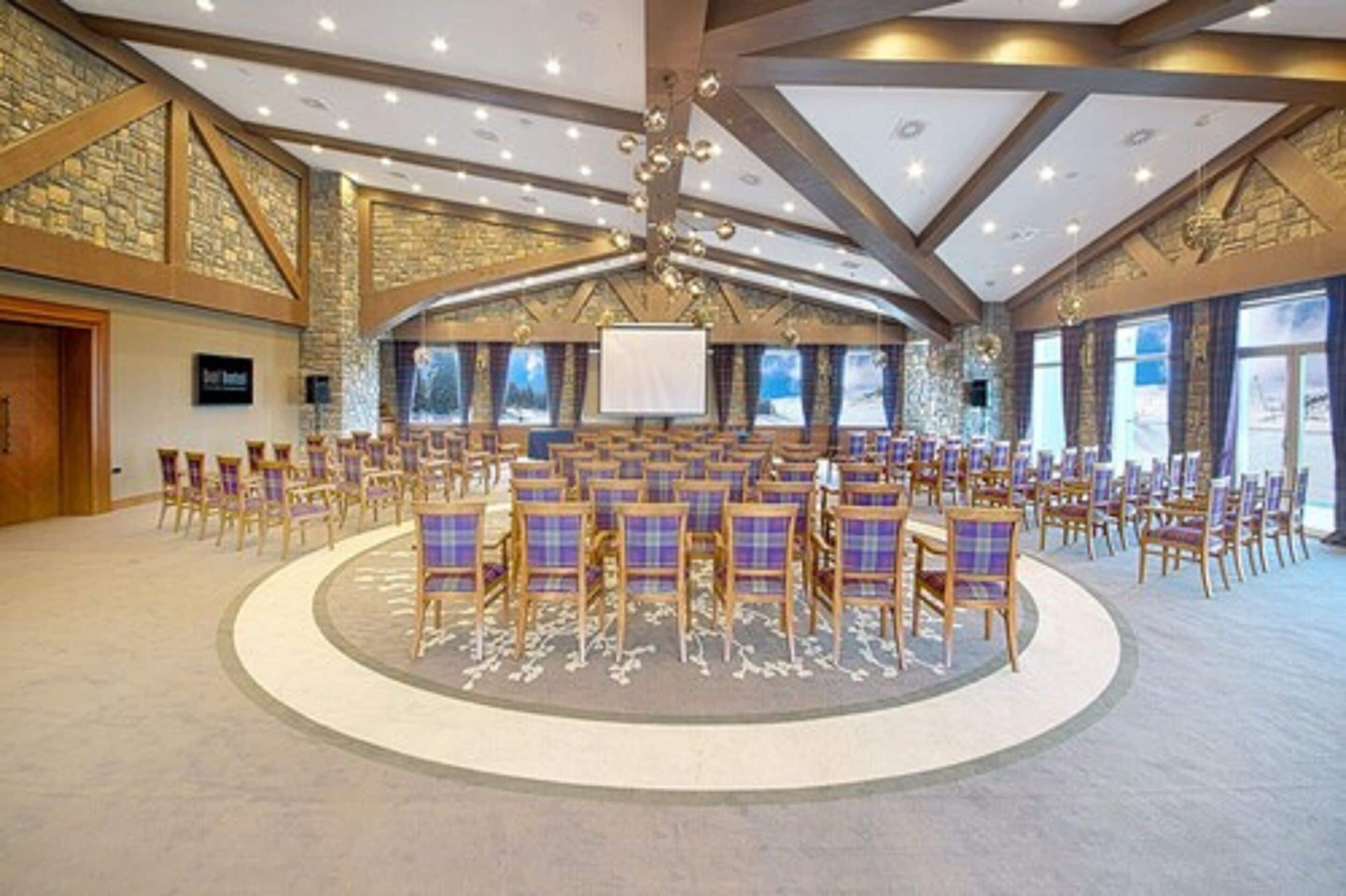 Une grande salle de conférence avec des rangées de chaises, une scène centrale et un mur en pierre en arrière-plan.