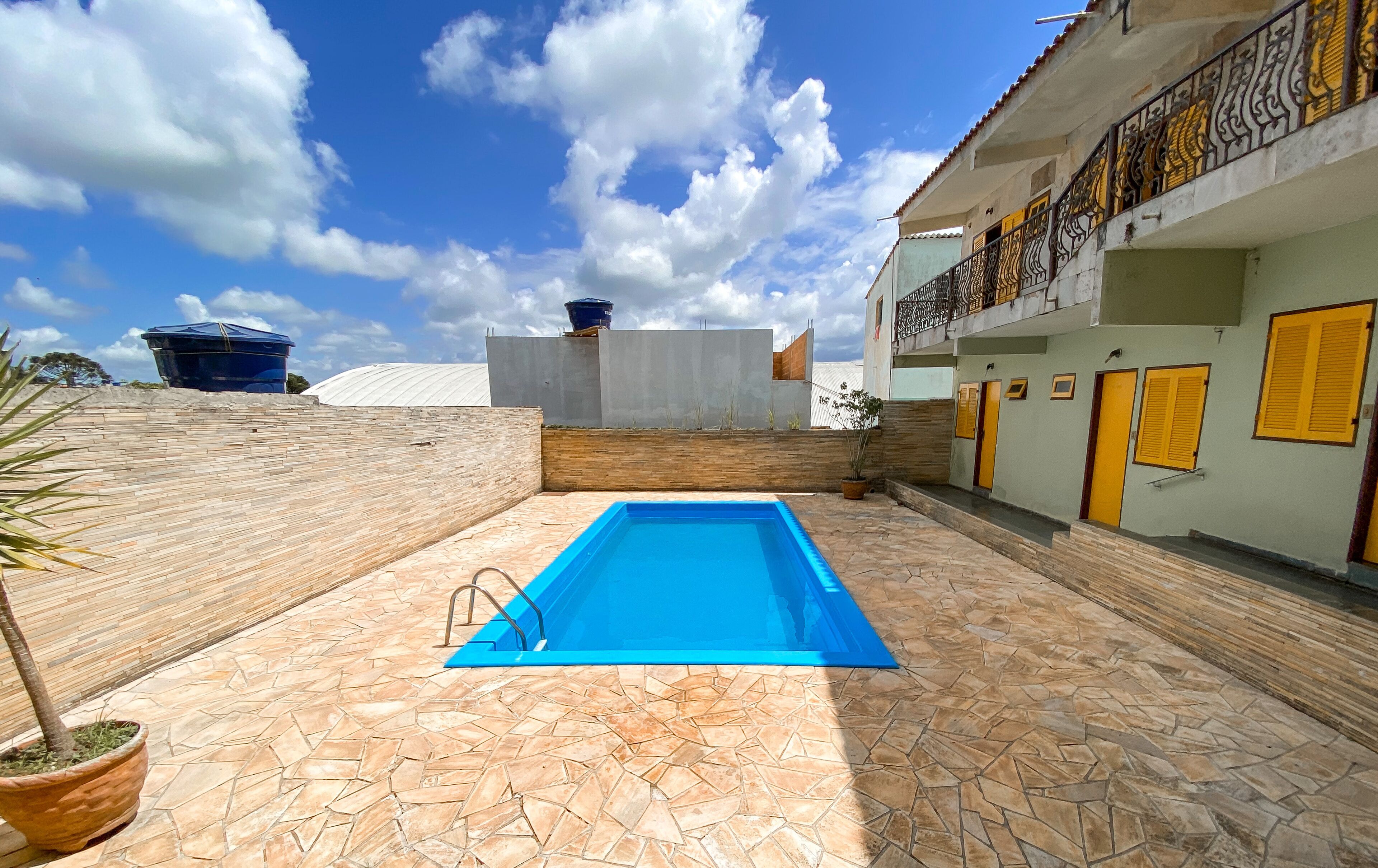 Una piscina con un patio de piedra, un edificio con puertas amarillas y un cielo azul despejado.