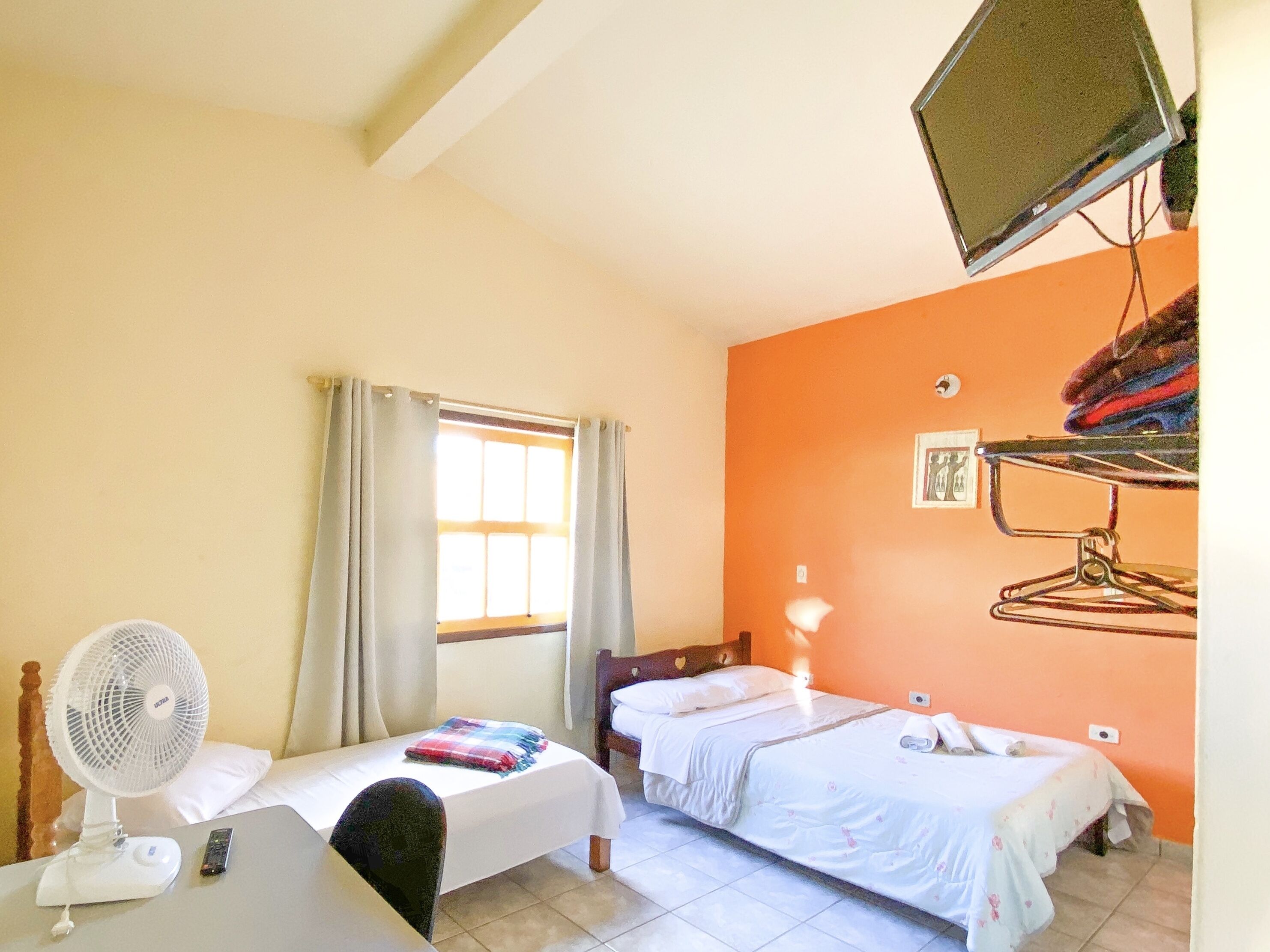 A hotel room with two beds, a desk with a fan, a window with curtains, and a wall-mounted TV.