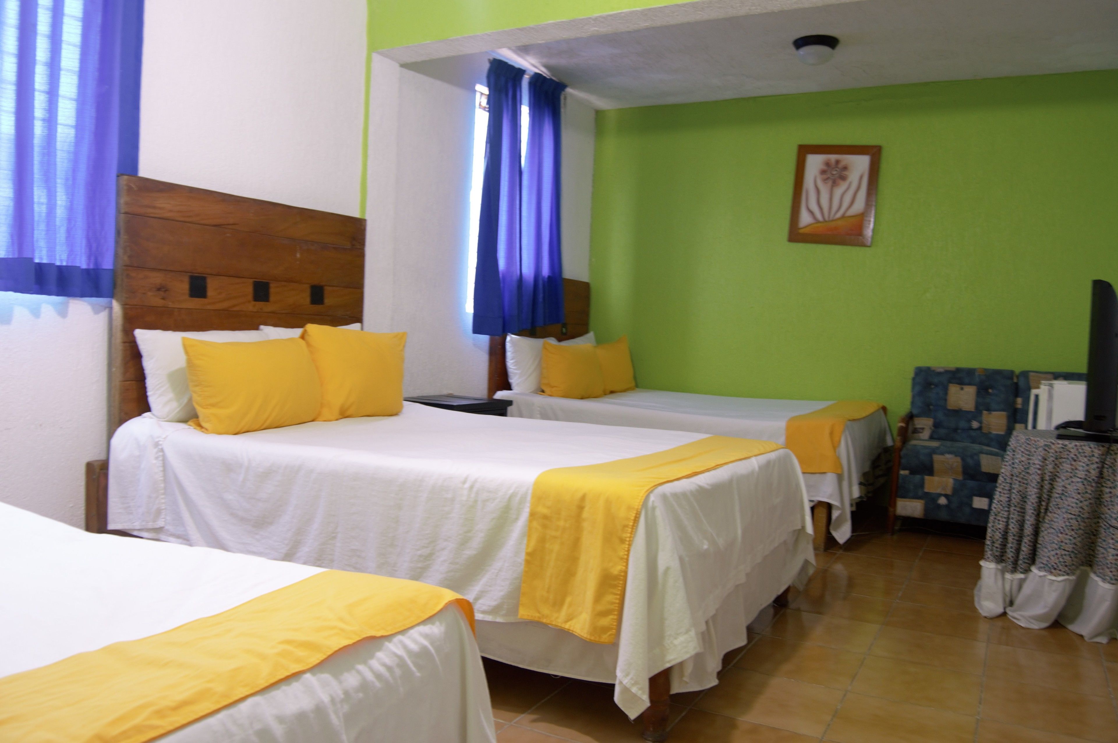 A hotel room with three beds, each with white bedding and yellow accents. The room has green walls and a wooden headboard.