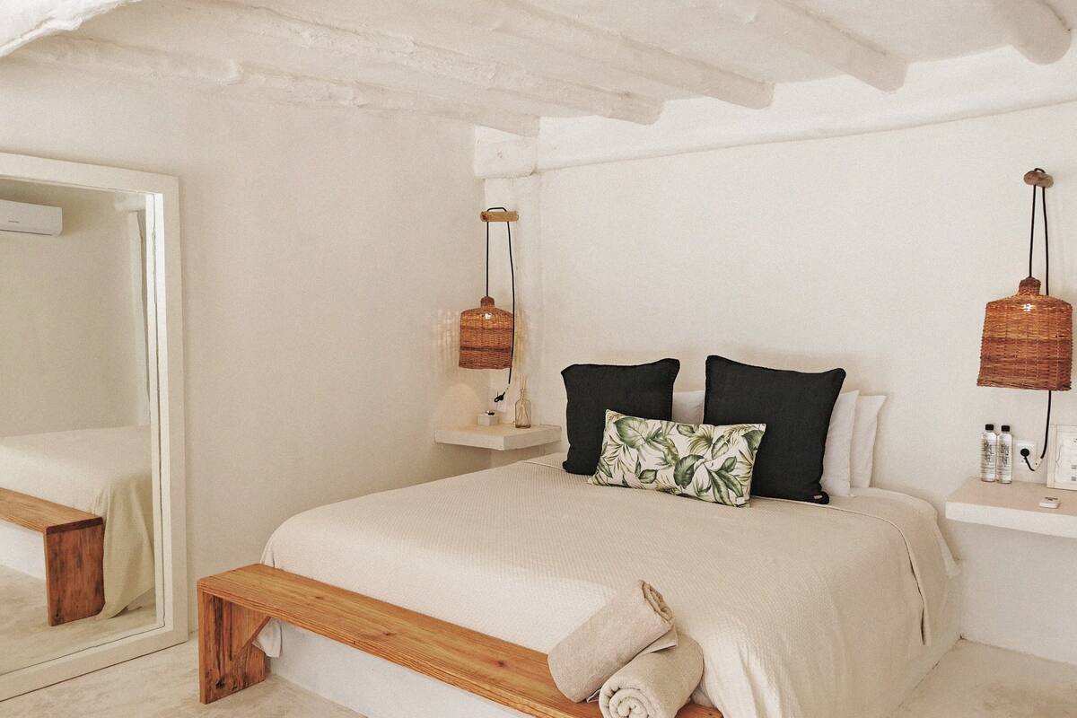 Un dormitorio con estructura de cama de madera, un espejo grande, dos mesitas de noche con lámparas de mimbre y un banco al pie de la cama.