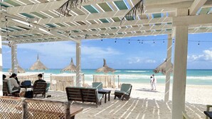 A beachside patio with wooden chairs and tables, thatched umbrellas, and a view of the ocean. - Coco Unlimited (Tulum)