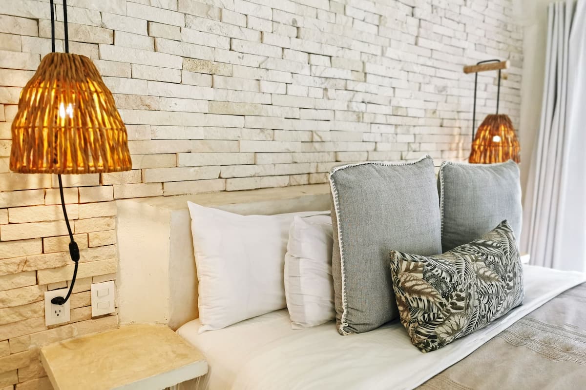 Un dormitorio con pared de ladrillo, mesita de noche de madera, cama con ropa de cama blanca y gris, y dos lámparas colgantes.