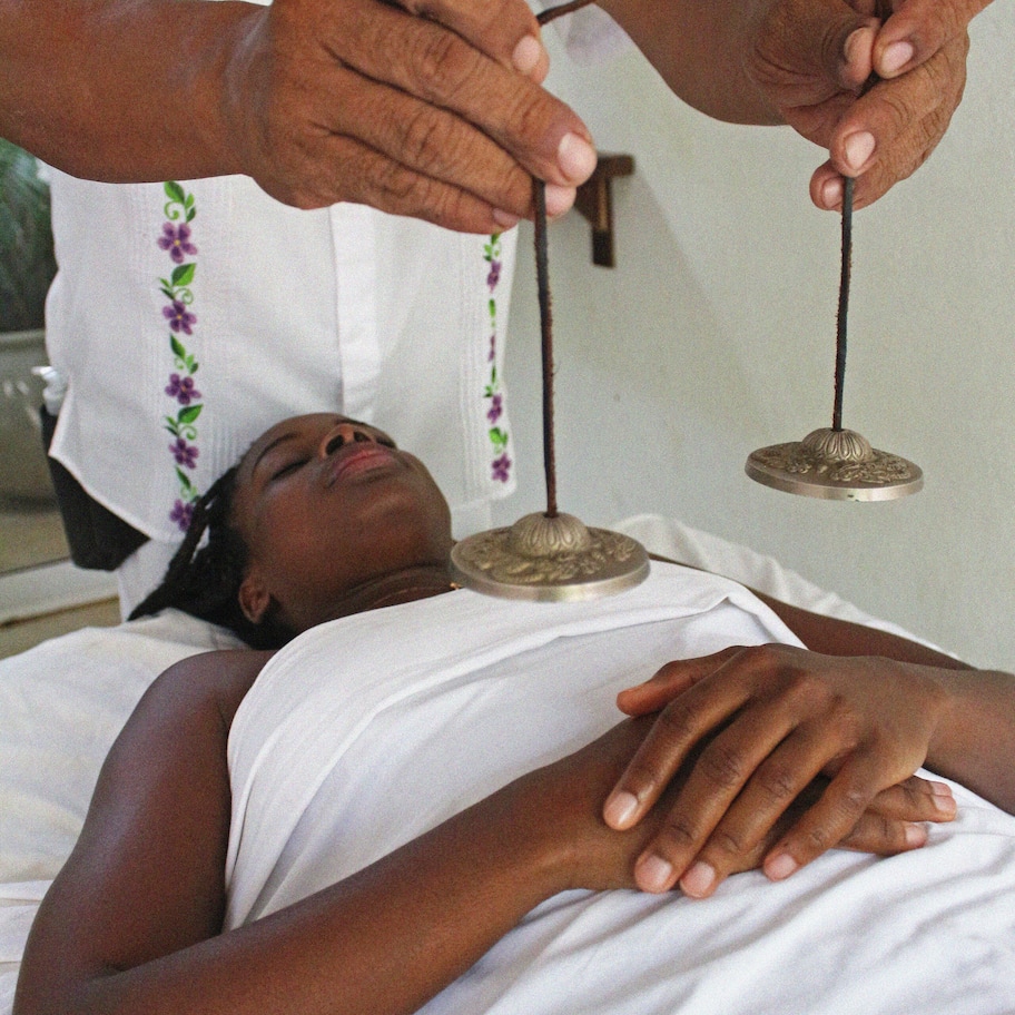 A person lying on a bed with a white sheet, being held by another person wearing a white shirt with floral embroidery.