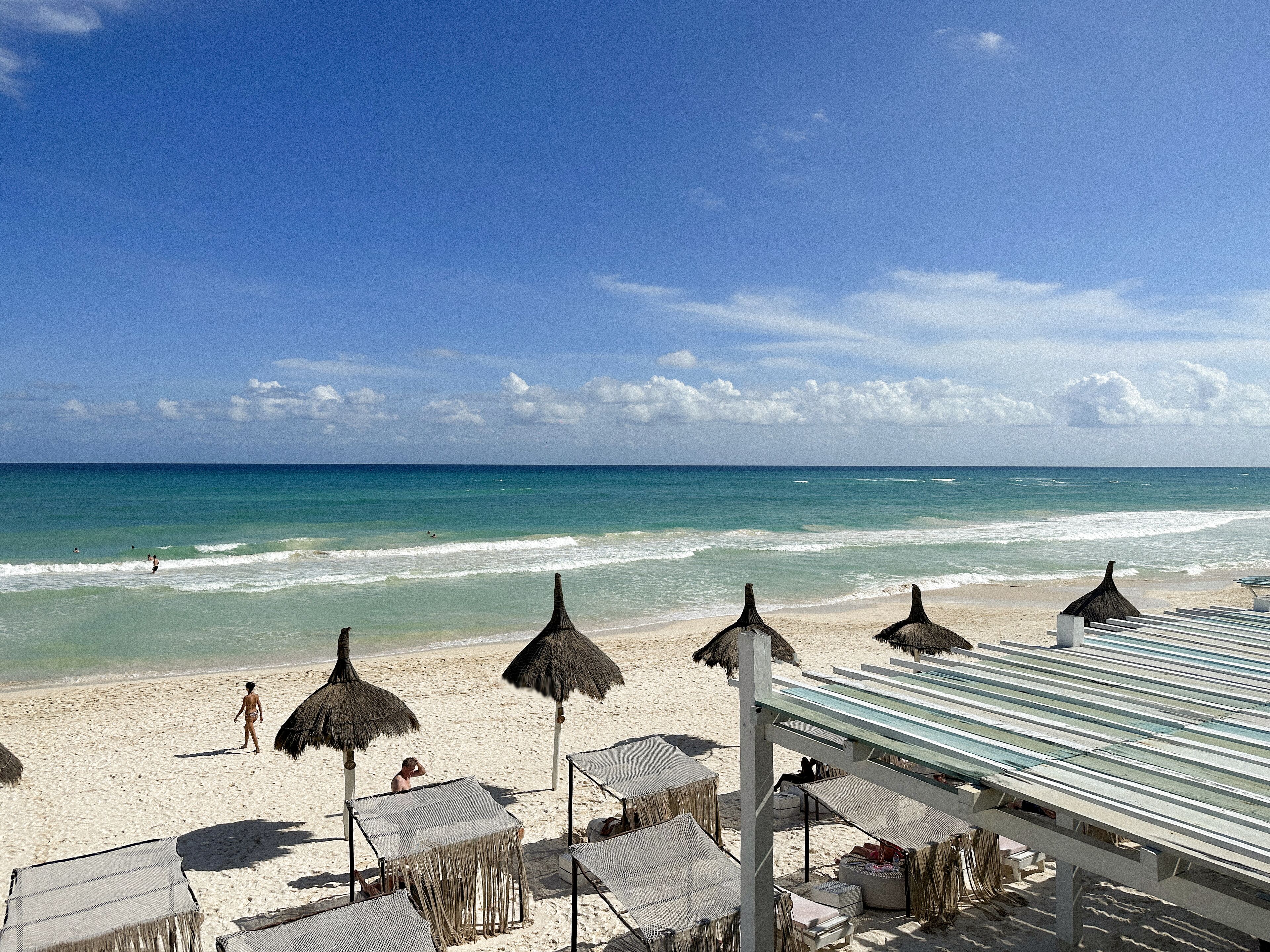 A beach with thatched umbrellas and lounge chairs, a clear sky, and the ocean.