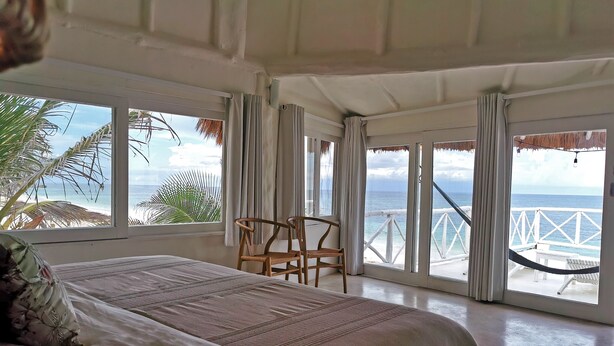 A bedroom with a large bed, a chair, and a view of the ocean through large windows. - Coco Unlimited (Tulum)