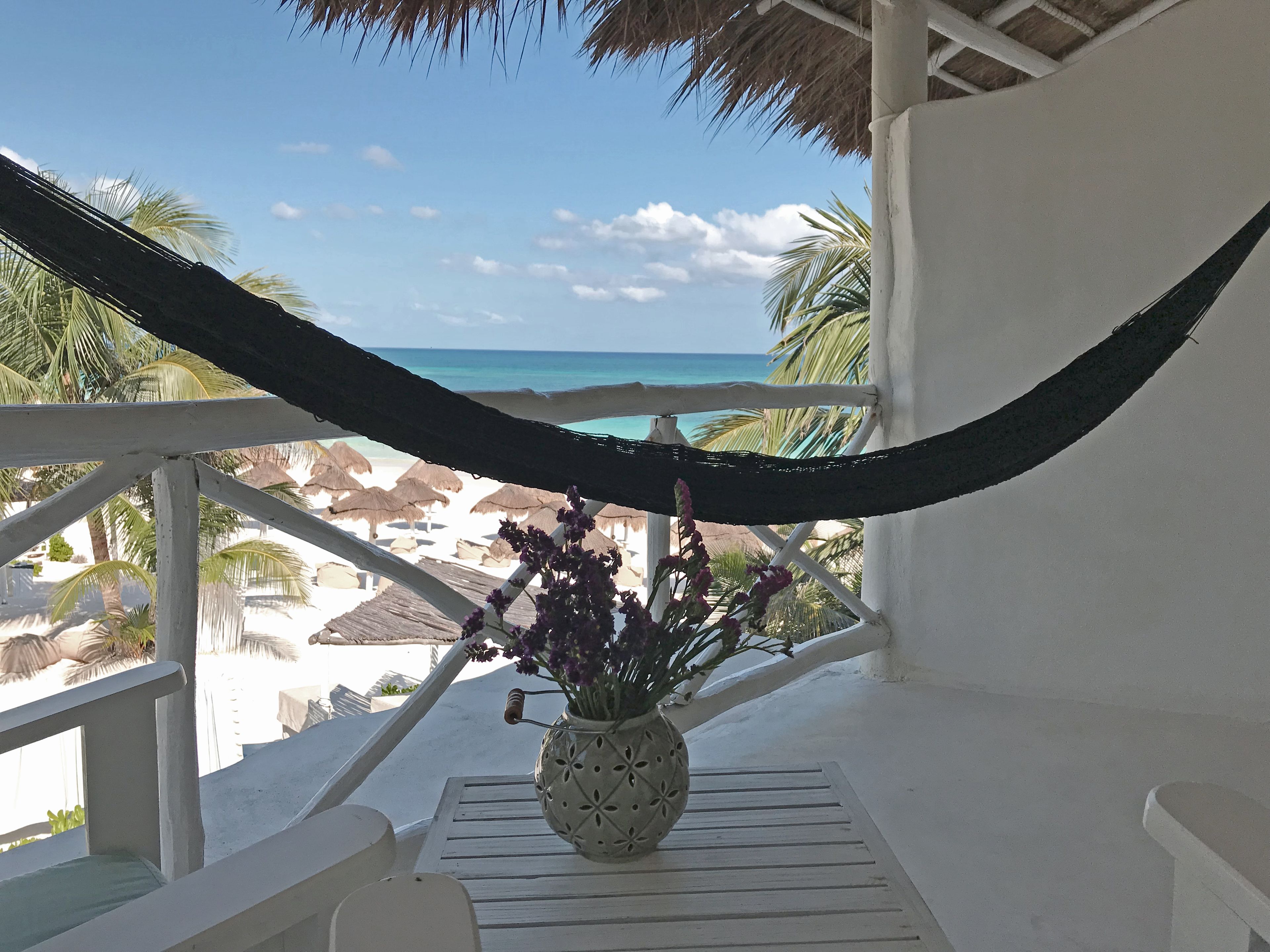 A hammock on a balcony overlooking a beach with thatched umbrellas and palm trees.