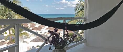 A hammock on a balcony overlooking a beach with thatched umbrellas and palm trees.