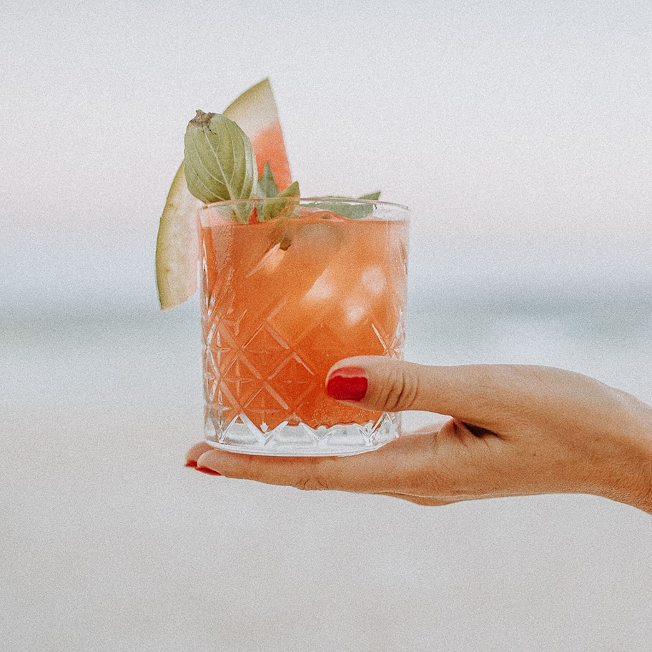 A hand holding a glass with a drink garnished with a mint leaf and a slice of lime, set against a blurred background.