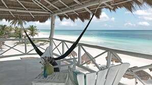 A beachside lounge with a thatched roof, white wooden chairs, and a table with a vase of flowers. - Coco Unlimited (Tulum)