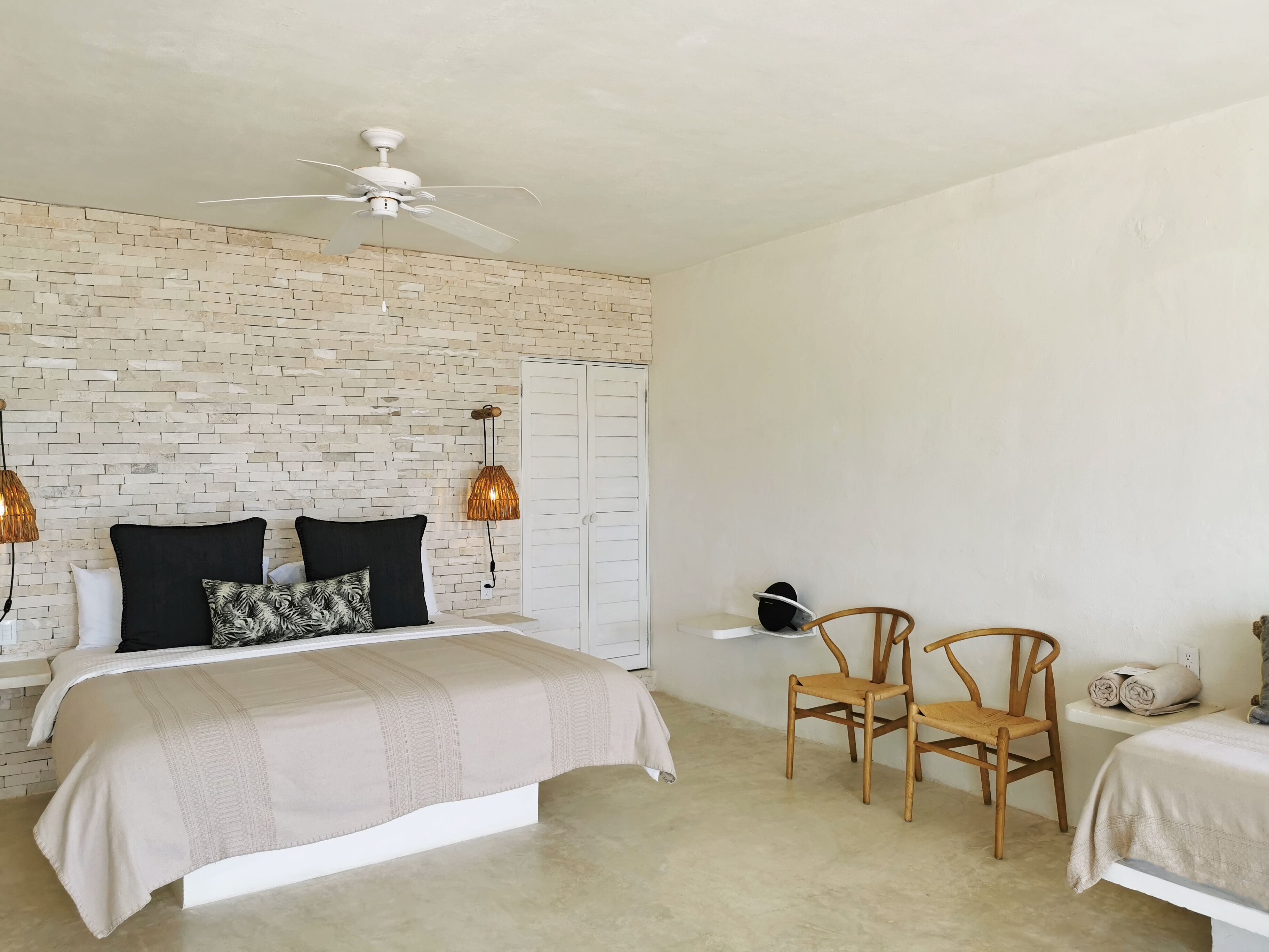 Una camera da letto con parete a motivo di mattoni, un letto con copripiumino beige, due sedie in legno, una ventola a soffitto e un'armadio bianco.