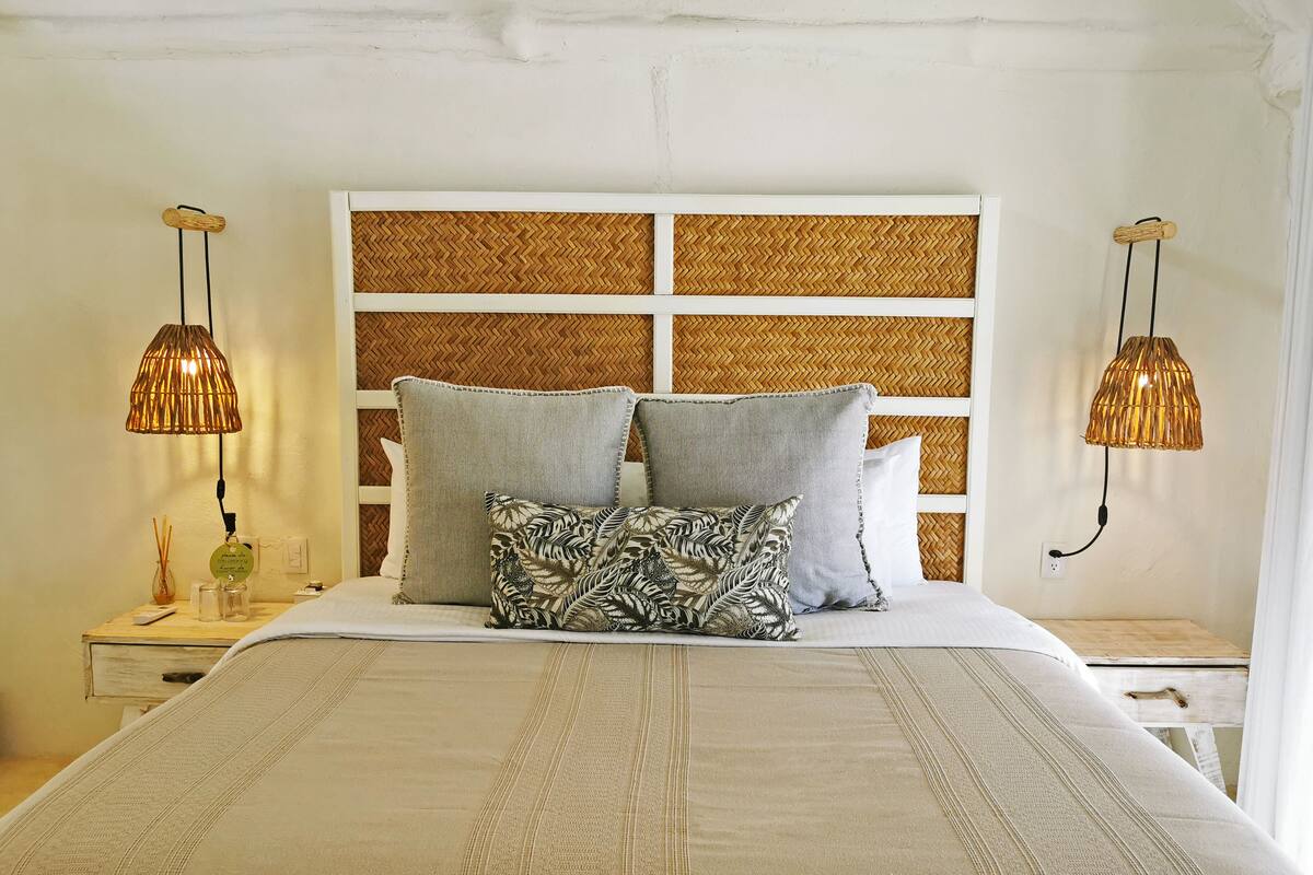 Una cama bien hecha con una colcha a rayas, dos almohadas y un cabecero tejido. Hay dos mesitas de noche con lámparas a ambos lados de la cama.