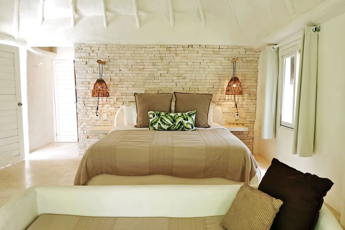 Un dormitorio con pared de ladrillo, una cama con cubrecama color crema, dos lámparas de pared y una ventana con cortinas.