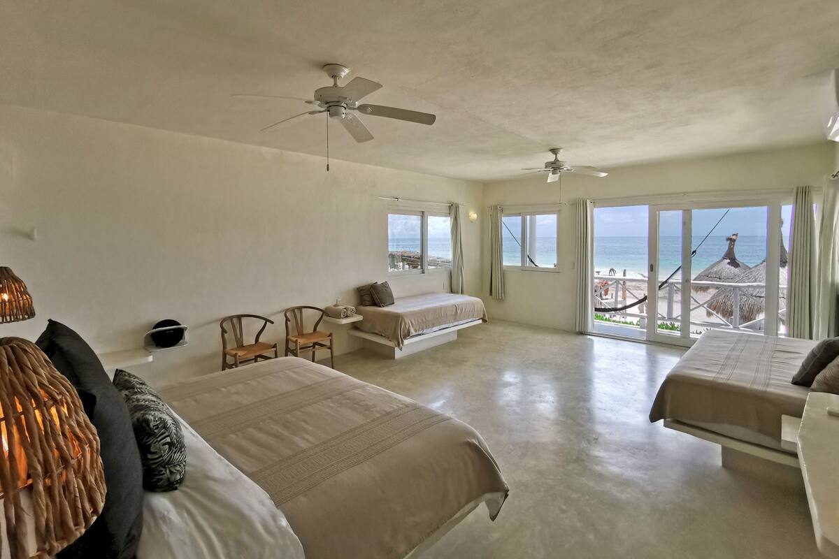 Un dormitorio amplio con dos camas, un ventilador de techo y un balcón con vistas a la playa.