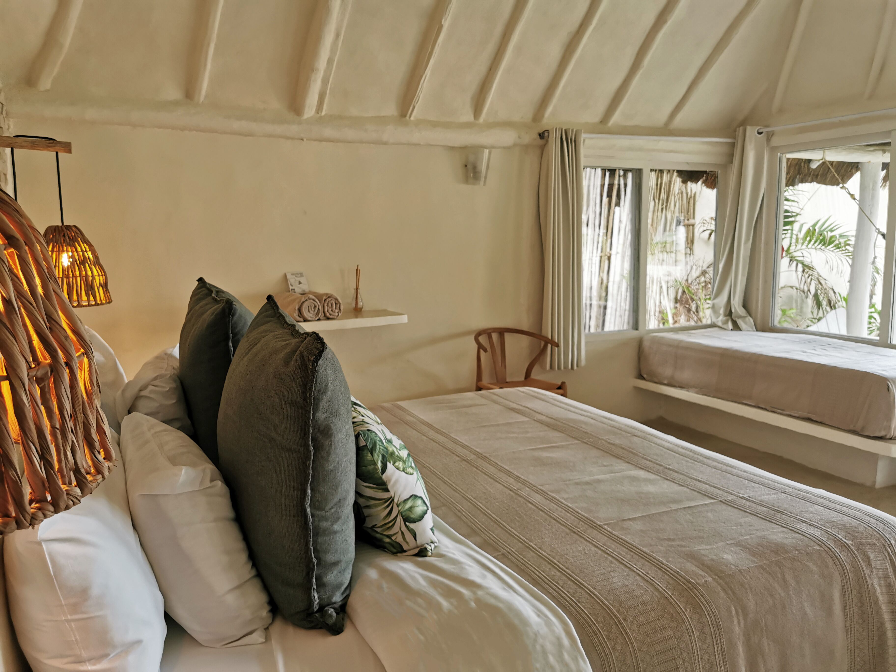 A bedroom with a bed, pillows, a hanging light fixture, a window with curtains, and a shelf with towels.
