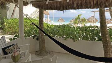 A beach view from a covered area with a hammock, white chairs, and thatched umbrellas.