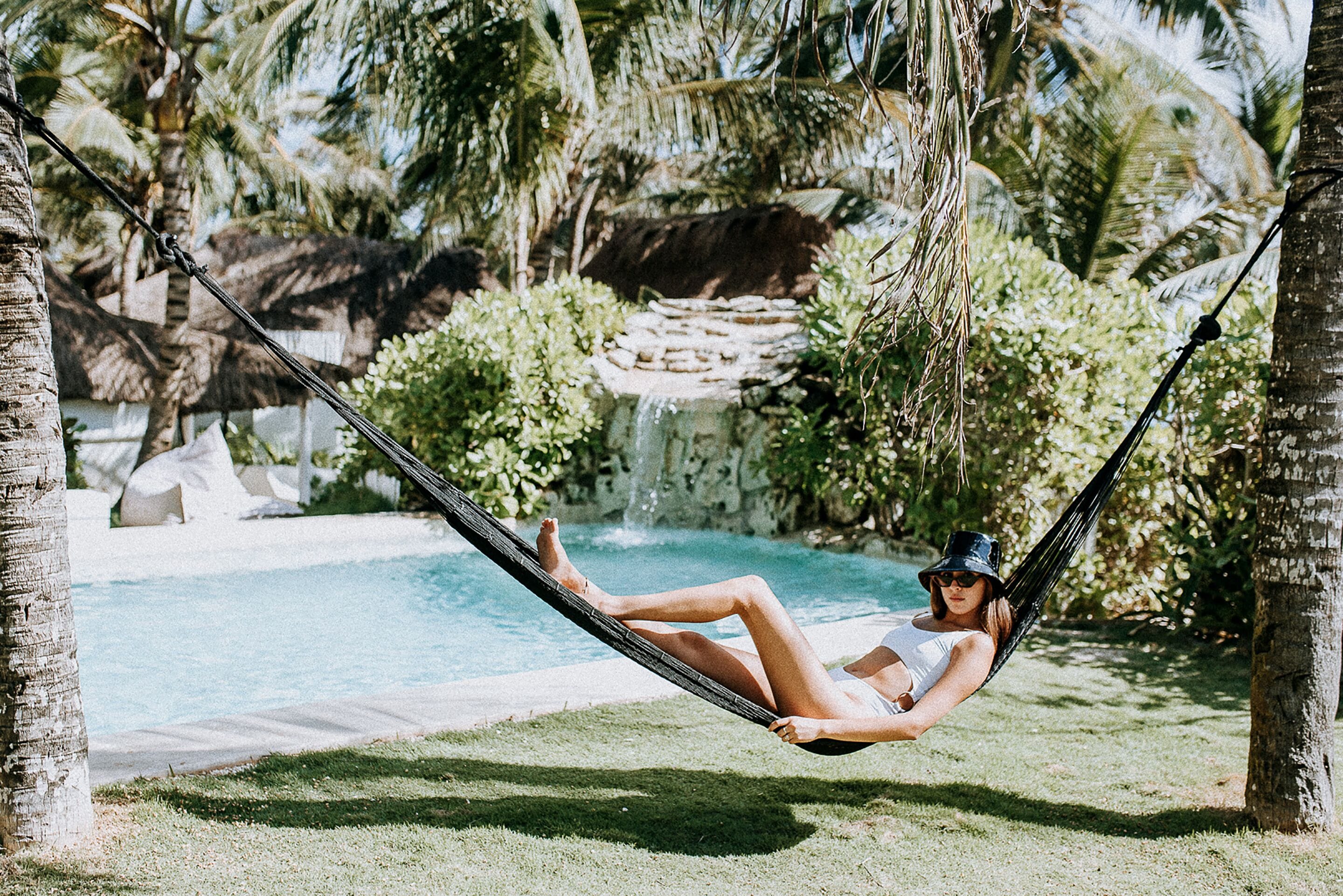 A hammock over a pool with palm trees and a waterfall in the background.