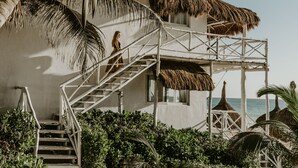 A beach house with a staircase leading to a balcony, surrounded by palm trees and a thatched roof structure. - Coco Unlimited (Tulum)