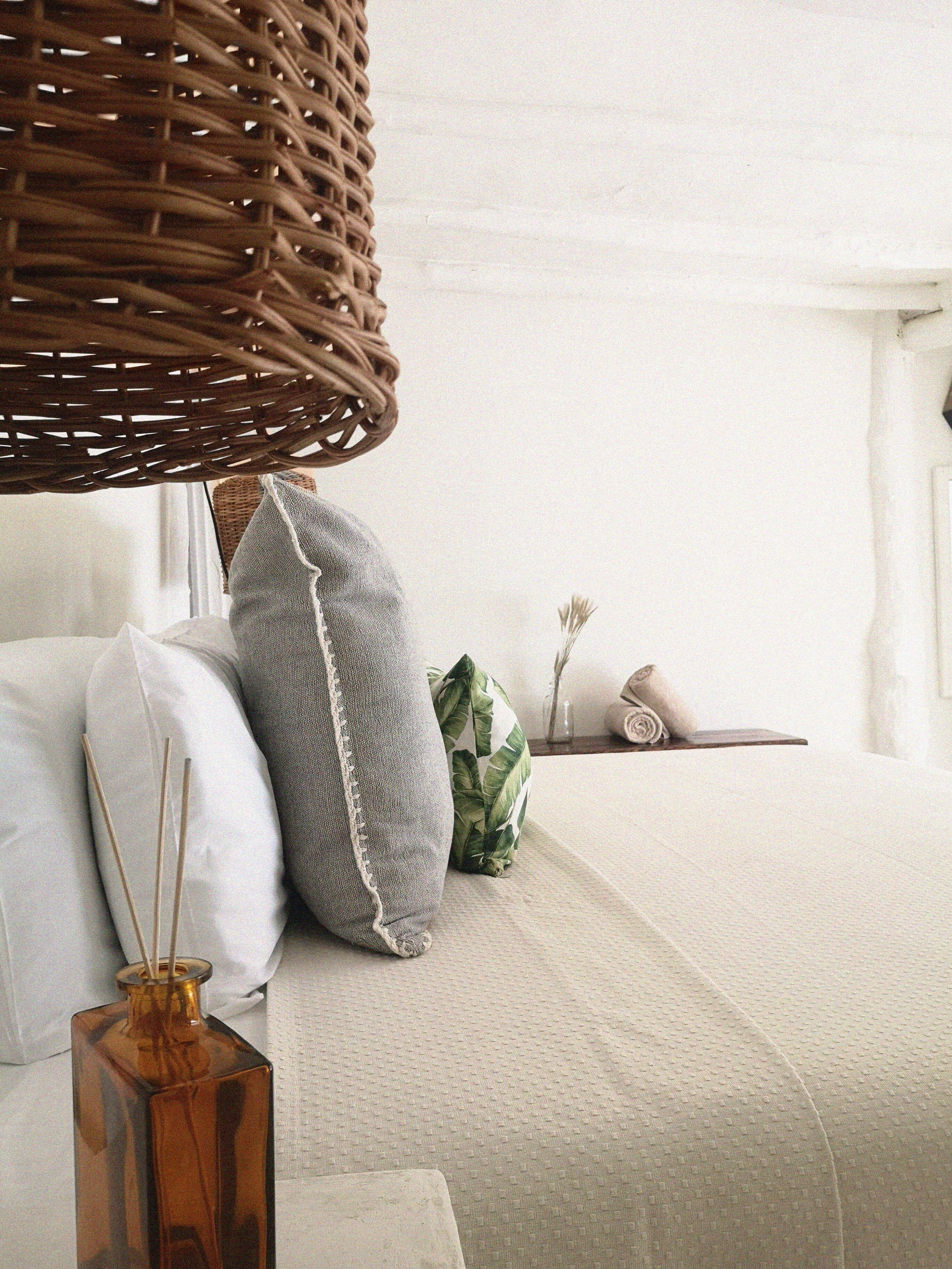 Un dormitorio ordenado con cama, almohadas, una cesta de mimbre y una mesita con un florero.